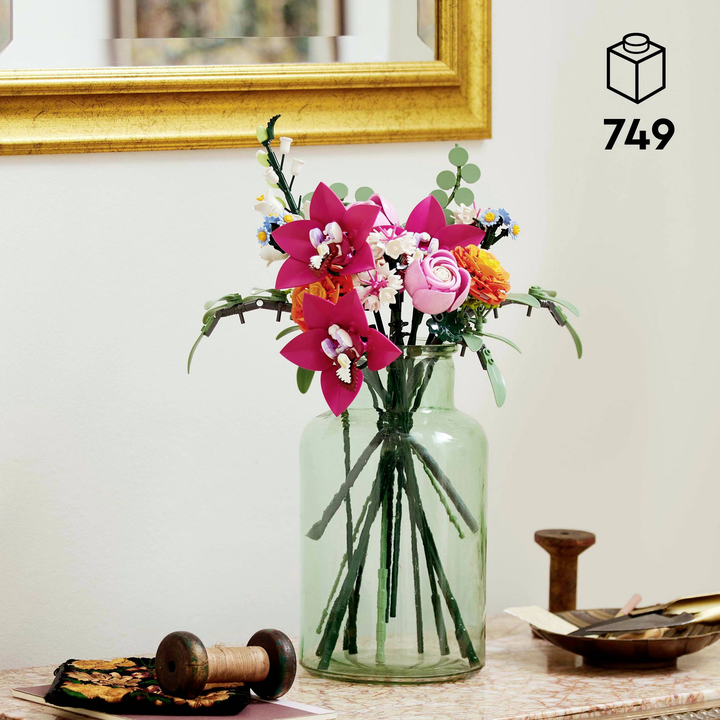 Un bouquet de fleurs en papier coloré dans un vase en verre est posé sur une table avec des accessoires de couture ; un miroir mural en arrière-plan.