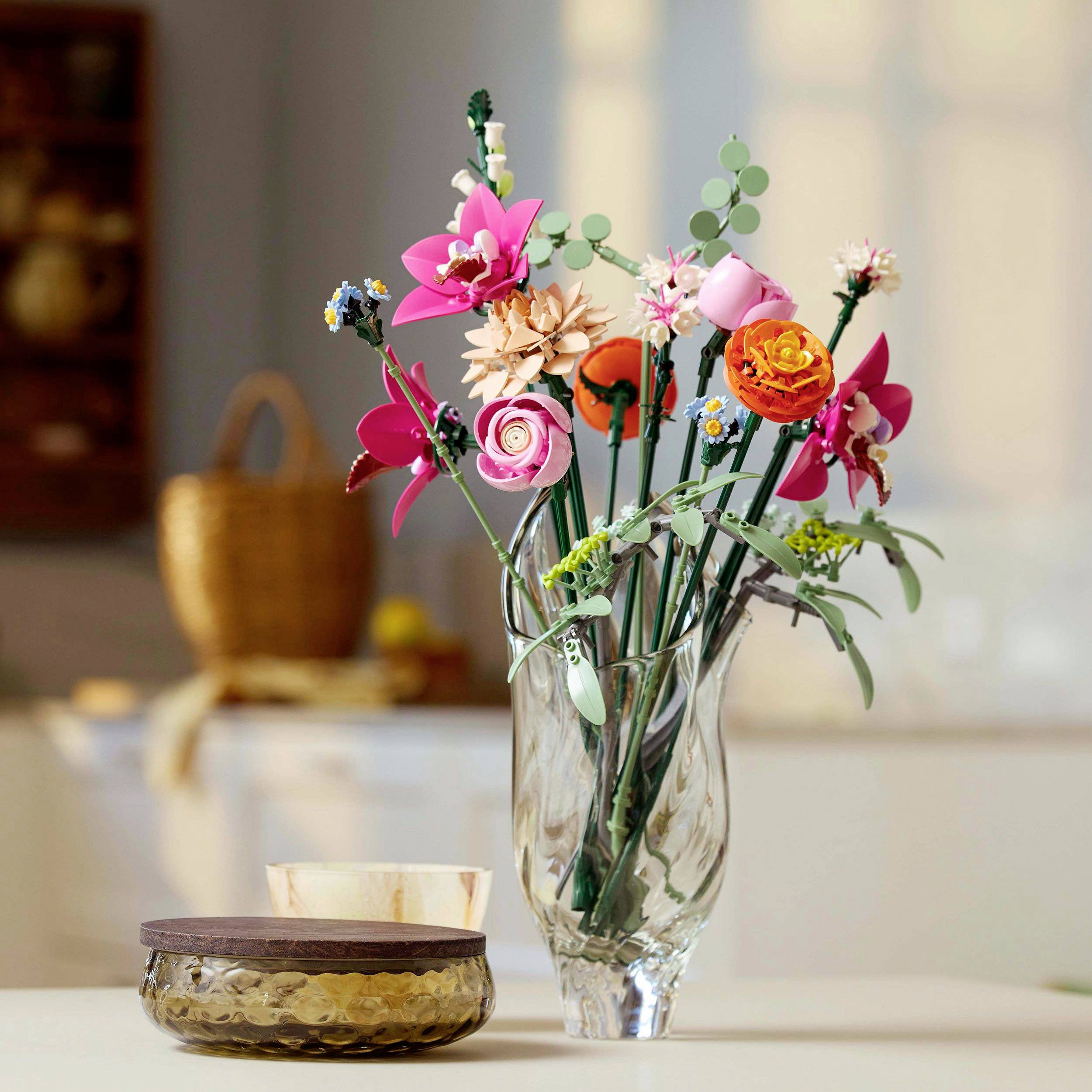 Un bouquet de fleurs dans une carafe en verre sur une table dans une cuisine lumineuse, entouré d'un bol et d'éléments de cuisine flous en arrière-plan.