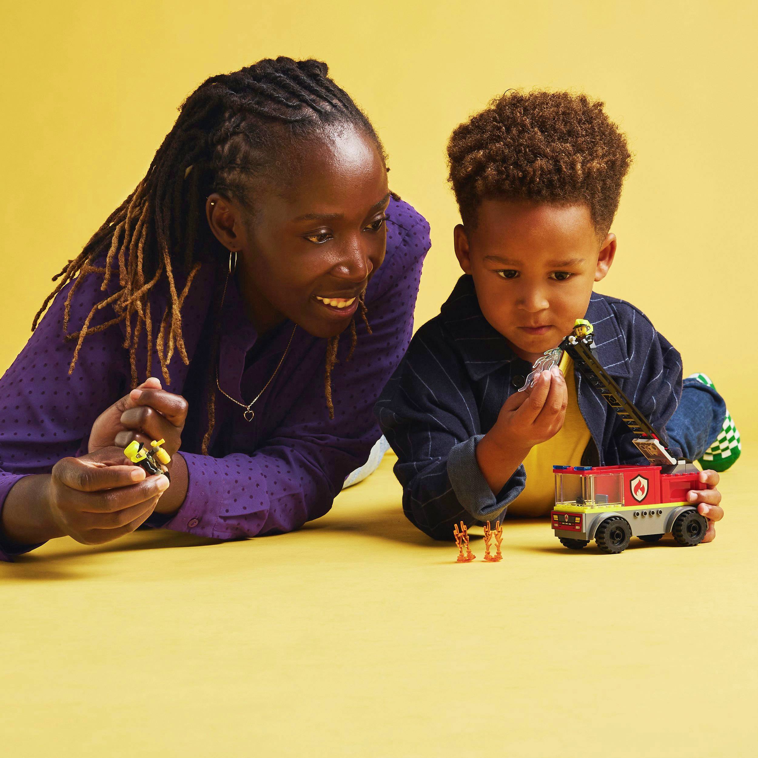 Une personne est allongée à côté d'un enfant par terre, tandis que l'enfant joue avec une voiture de pompiers en jouet et des figurines jaunes.
