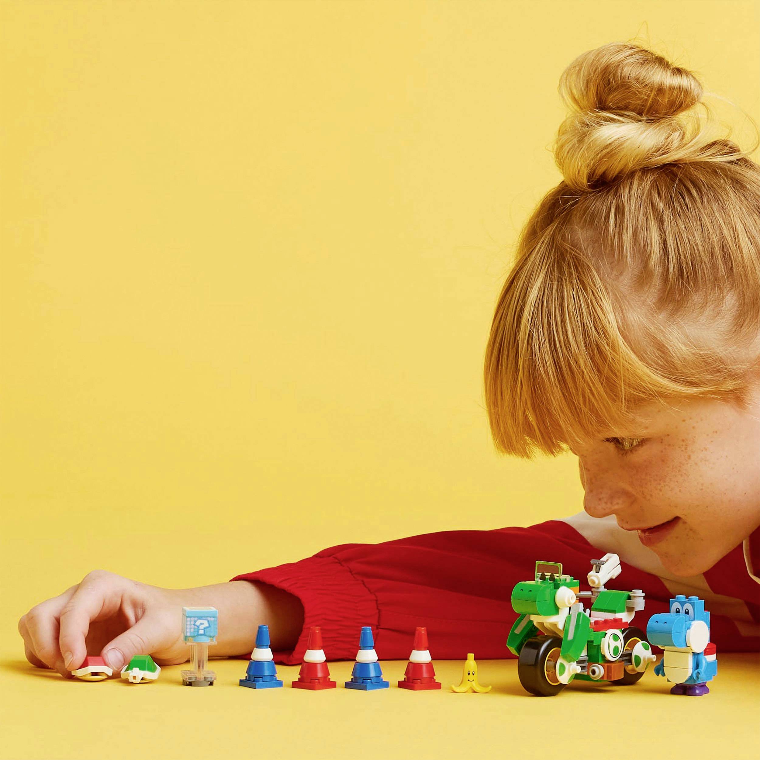 Un enfant aux cheveux roux est allongé sur le ventre et joue avec des figurines colorées et un véhicule jouet vert sur un fond jaune.
