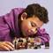 Un enfant joue avec des figurines Lego sur une table. La scène Lego représente une pièce avec un tableau et plusieurs personnages. L'enfant porte un sweat à capuche violet.