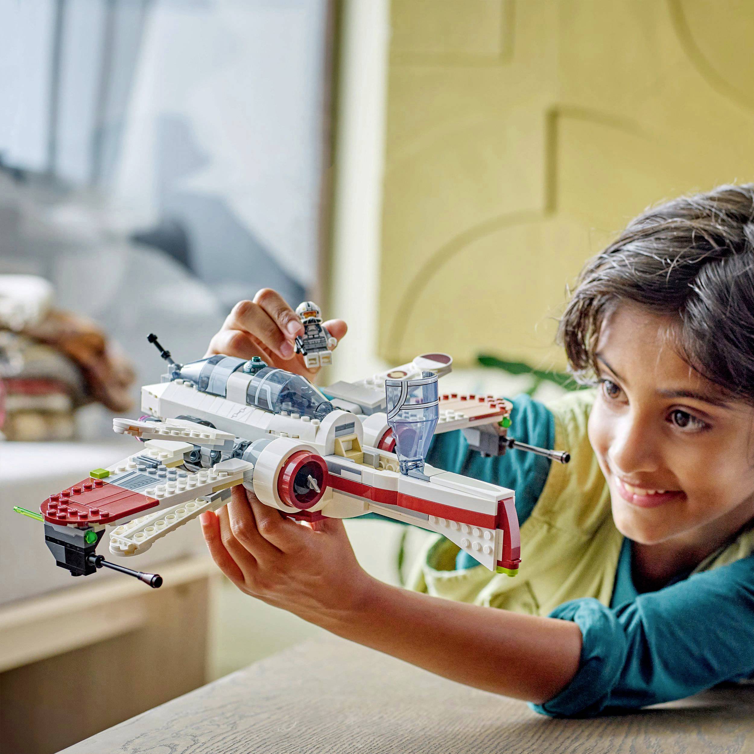 Un enfant joue en souriant avec un vaisseau spatial en Lego sur une table, avec des détails flous d'un intérieur en arrière-plan.