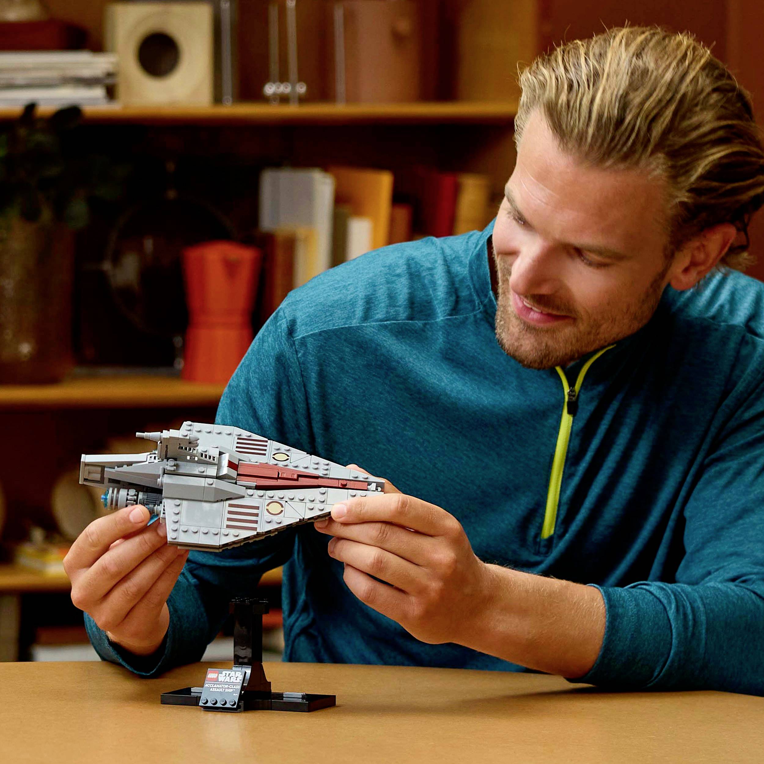 Un homme souriant tient un modèle de vaisseau spatial dans ses mains et l'observe. À l'arrière-plan, on voit des étagères remplies de livres et de décorations.