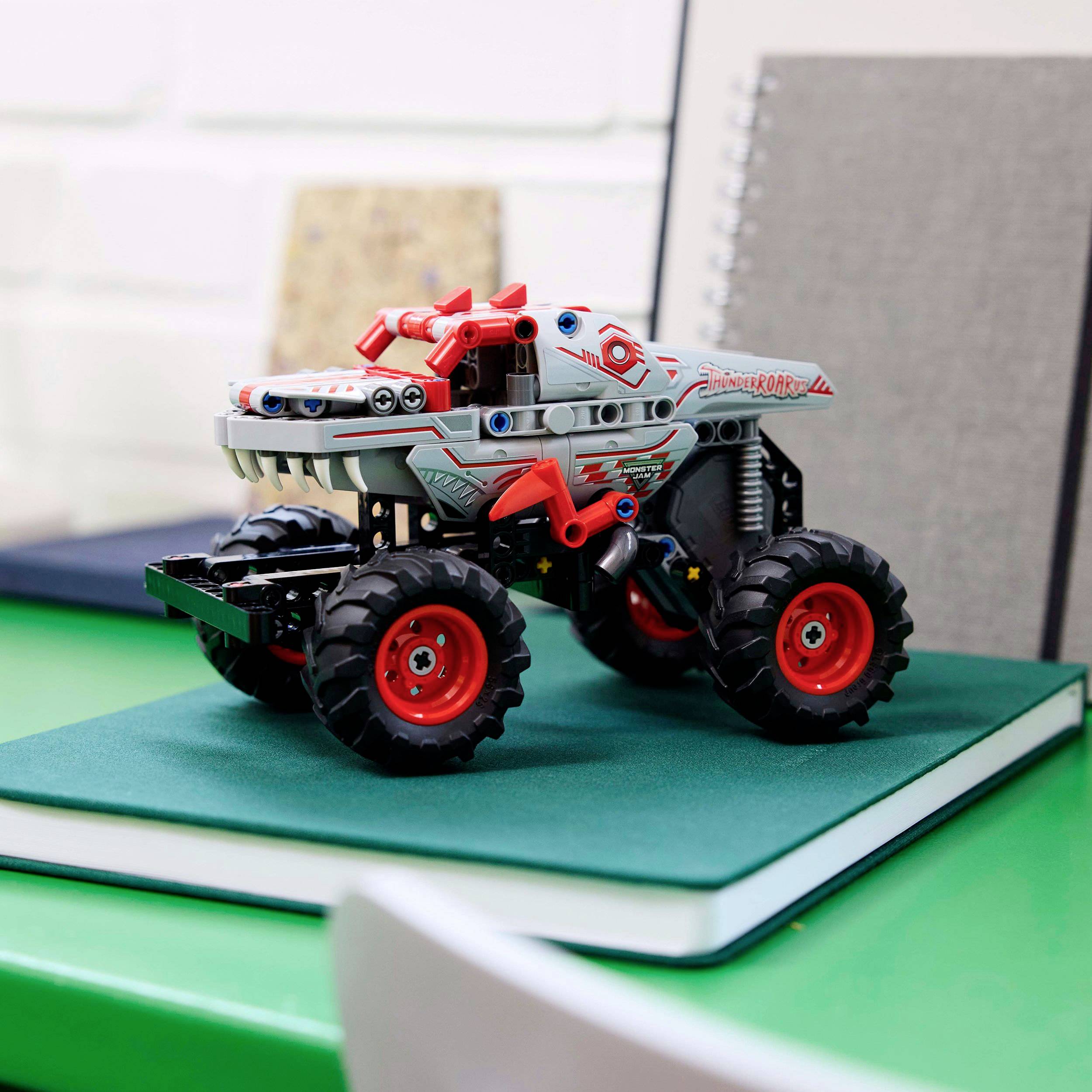 Une voiture miniature en forme de dinosaure est posée sur un carnet, sur une table verte. Des livres sont visibles en arrière-plan.