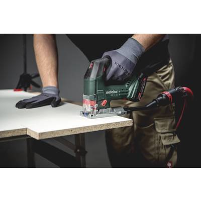 Une personne découpe une planche blanche en bois à l'aide d'une scie sauteuse. Elle porte des gants de travail et des vêtements de travail gris foncés.