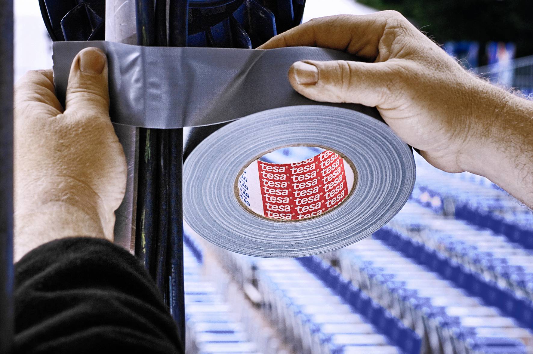 Deux mains fixent du ruban adhésif gris sur un poteau noir. En arrière-plan, des chaises bleues floues sont visibles.