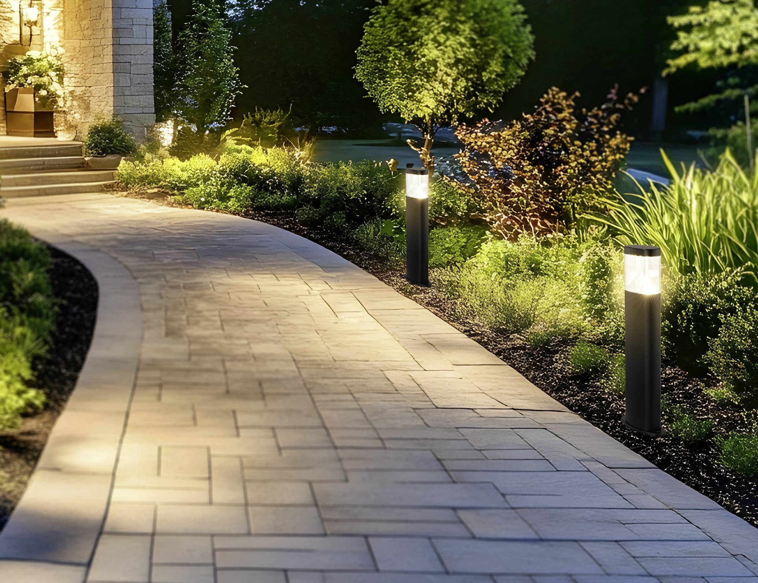 Un chemin de pierre courbe, éclairé par des luminaires de jardin, traverse un espace paysager verdoyant avec des arbustes luxuriants, menant à l'entrée d'une maison au crépuscule.