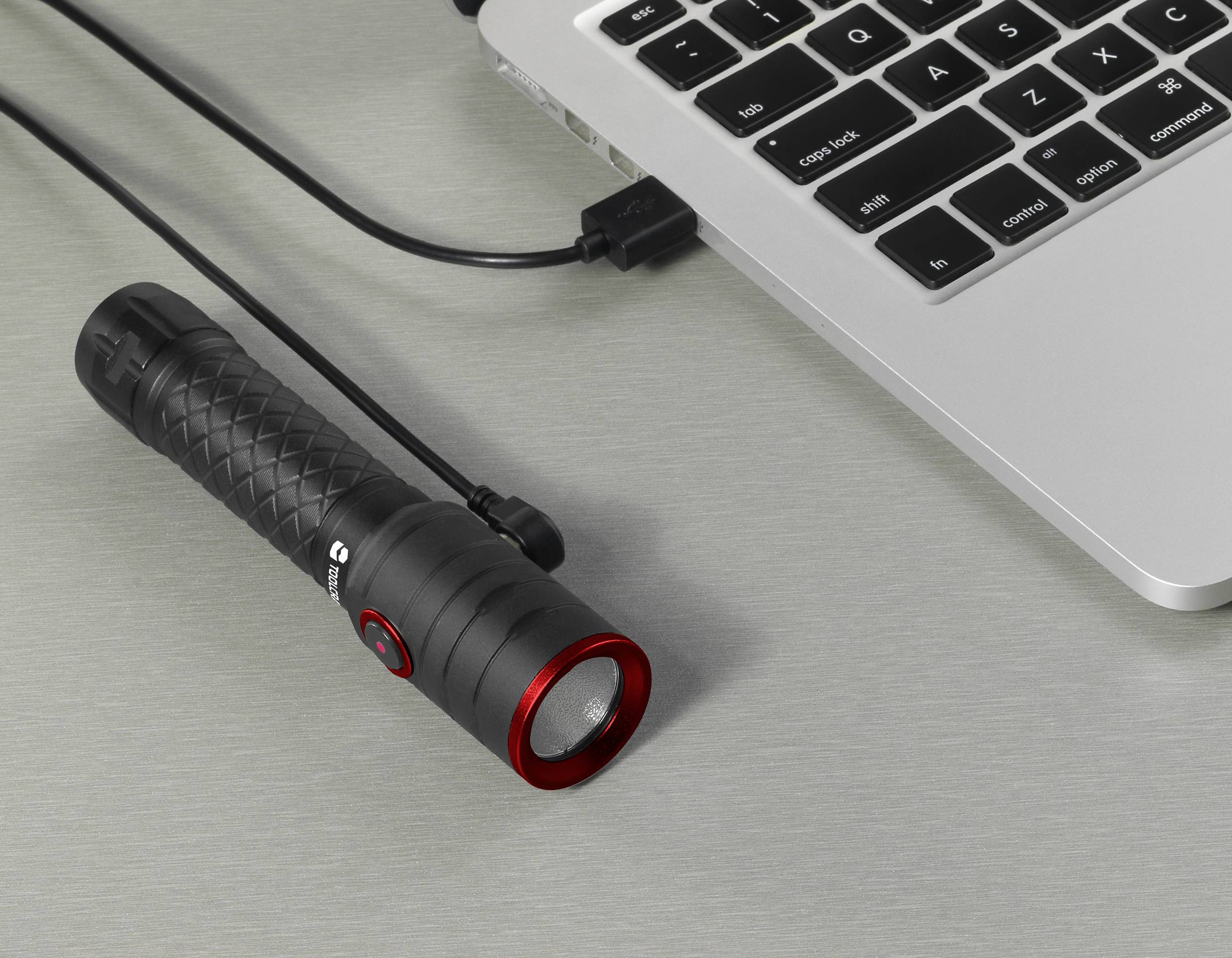 Une lampe de poche noire avec un bord rouge est connectée à un ordinateur portable via un câble USB. Elle est posée sur une table.