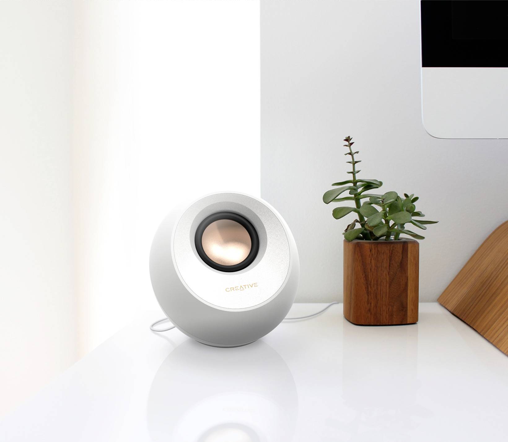 Enceinte blanche et ronde posée sur une table blanche à côté d'un pot en bois contenant une plante, avec une partie d'un moniteur visible.