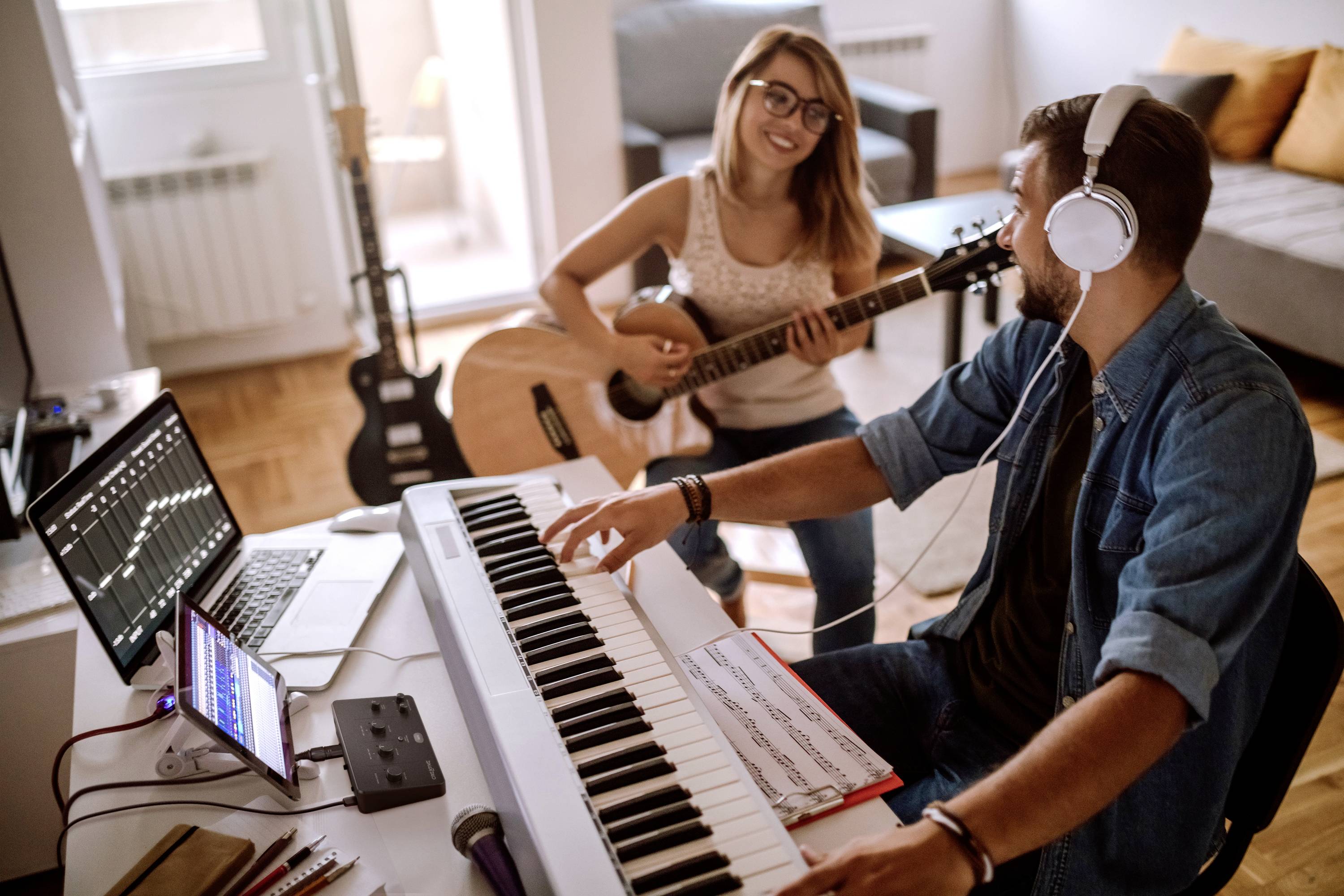 Un couple fait de la musique dans un salon bien éclairé. La femme joue de la guitare, l'homme, portant des écouteurs, joue du clavier. Des ordinateurs portables affichent des logiciels de musique.