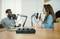 Deux personnes discutent dans un studio de podcast d'enregistrement, avec des microphones et une console de mixage audio posés sur une table en bois.