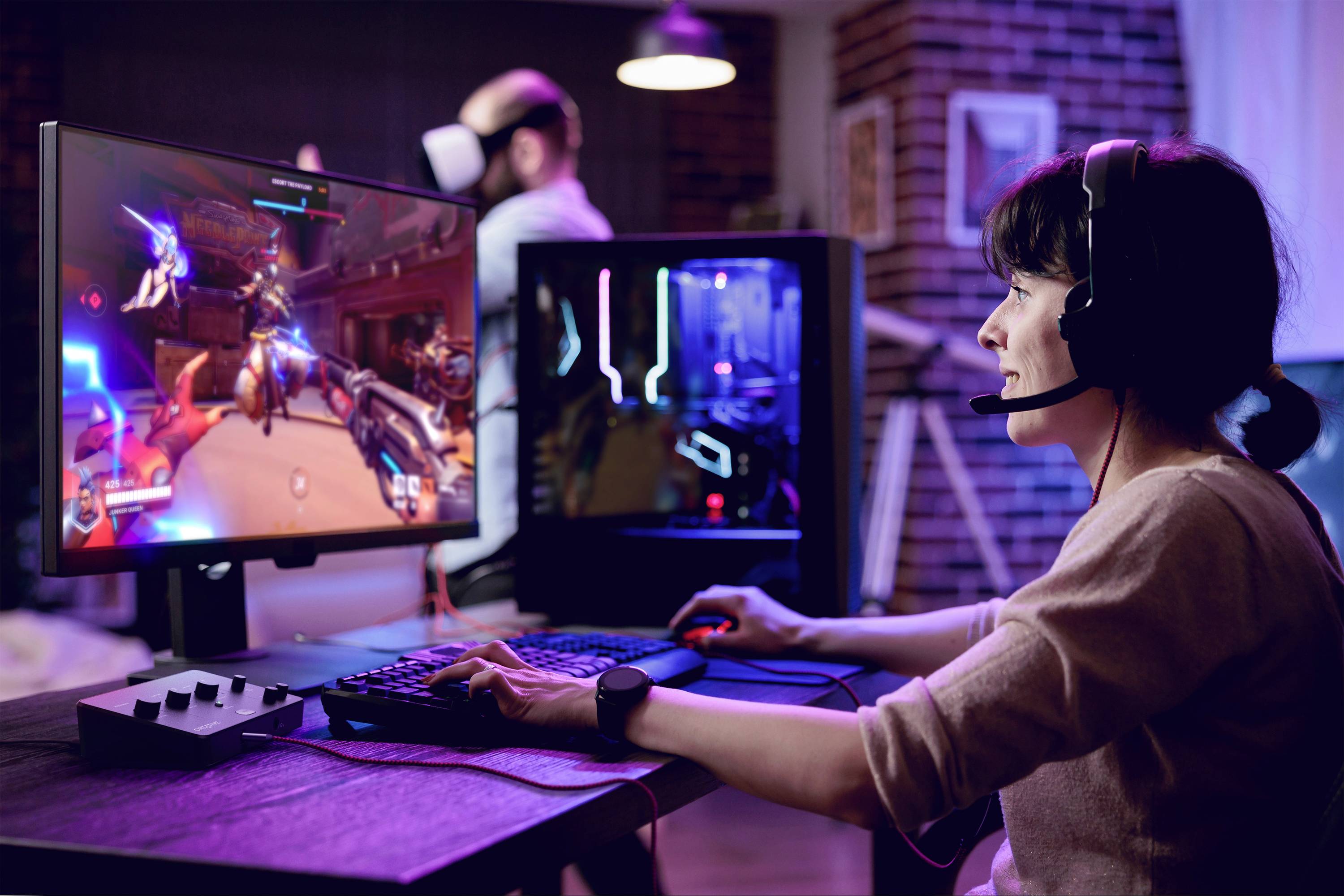 Femme portant un casque audio, jouant à un jeu vidéo sur PC, tandis qu'un homme porte un casque de réalité virtuelle en arrière-plan.