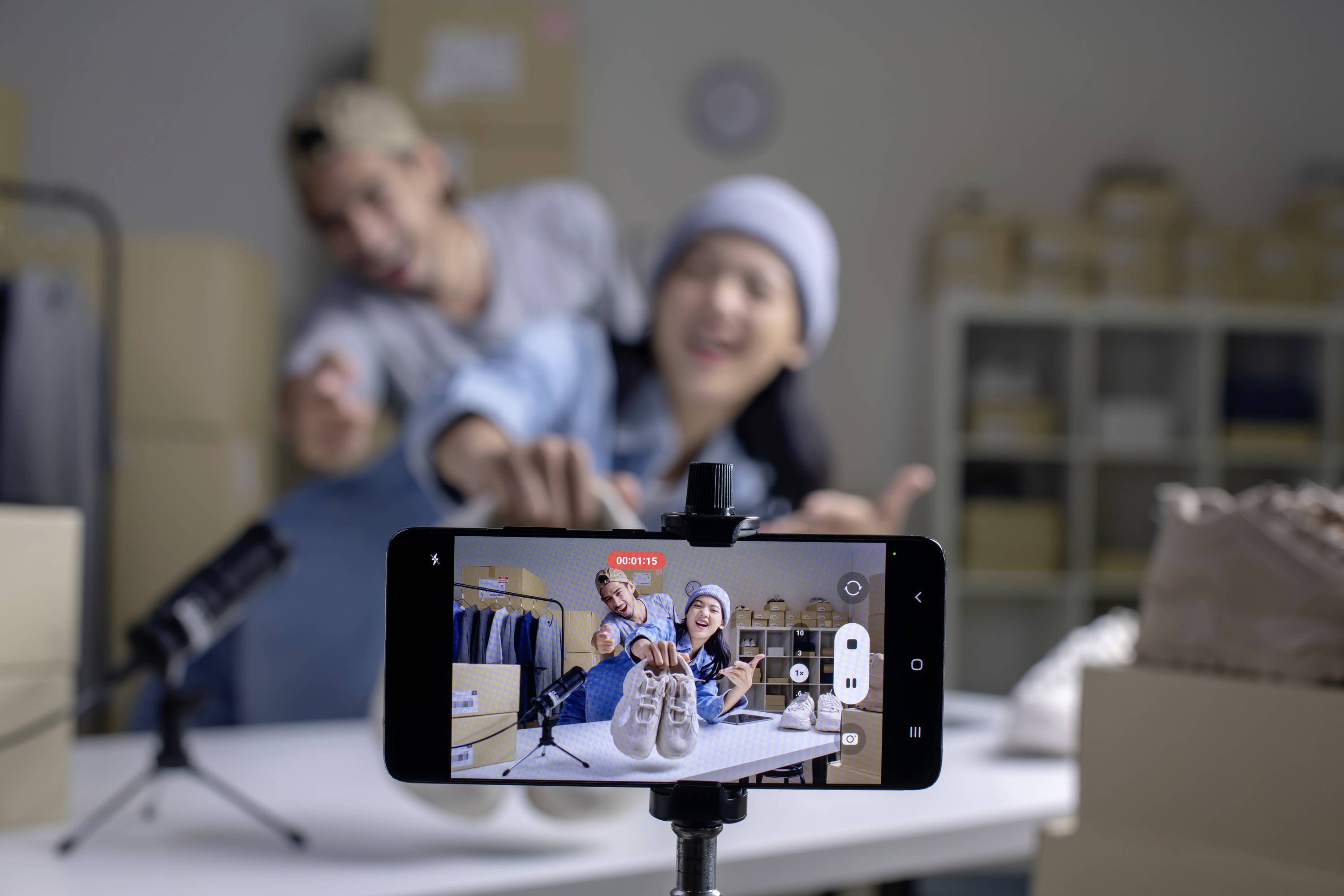 Un appareil photo de smartphone met au point sur des chaussures posées sur une table, tandis que deux personnes sourient et posent en arrière-plan.