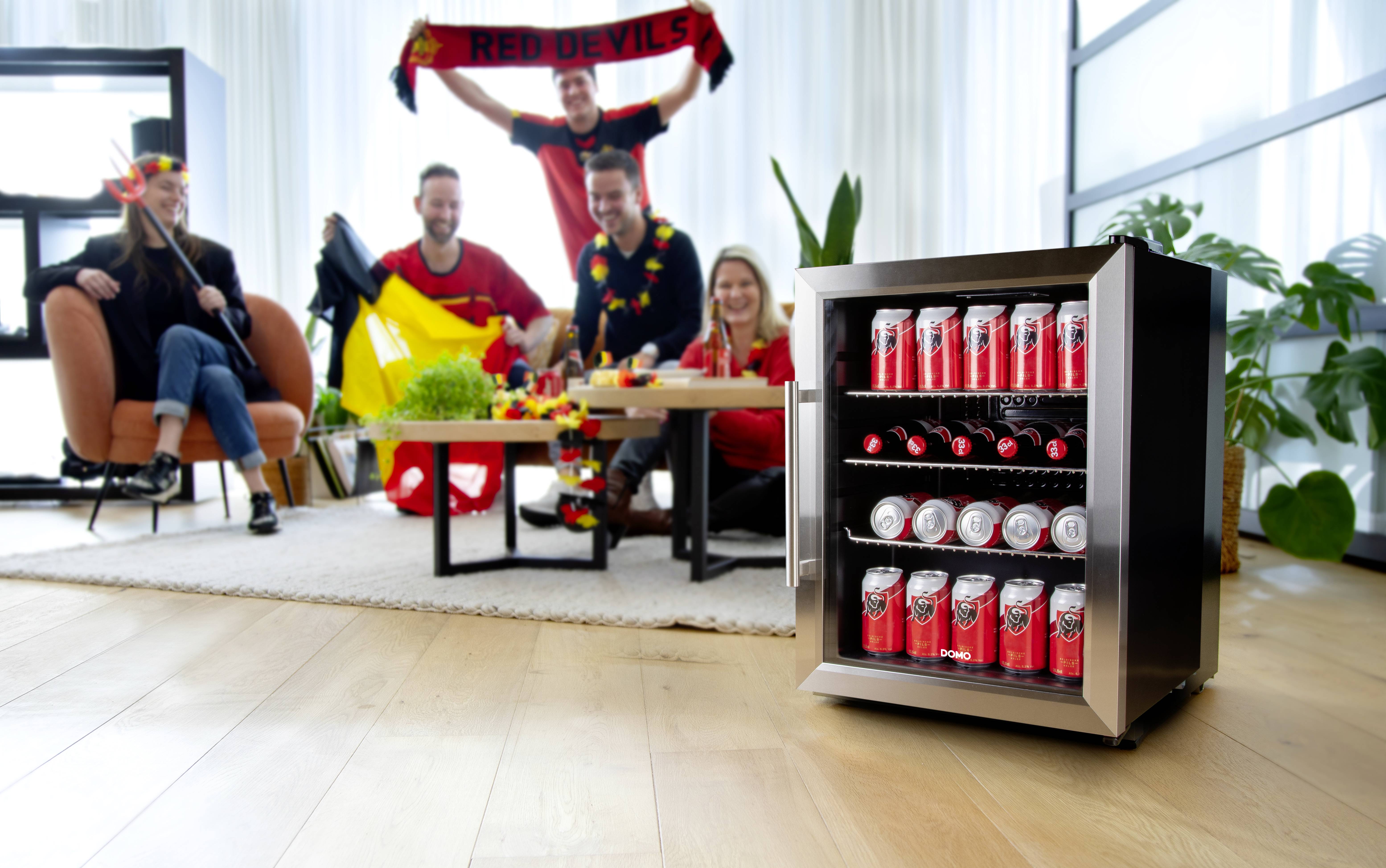 Un groupe de personnes portant des articles de fan est assis dans un salon. Au premier plan, un petit réfrigérateur est rempli de canettes de boissons.