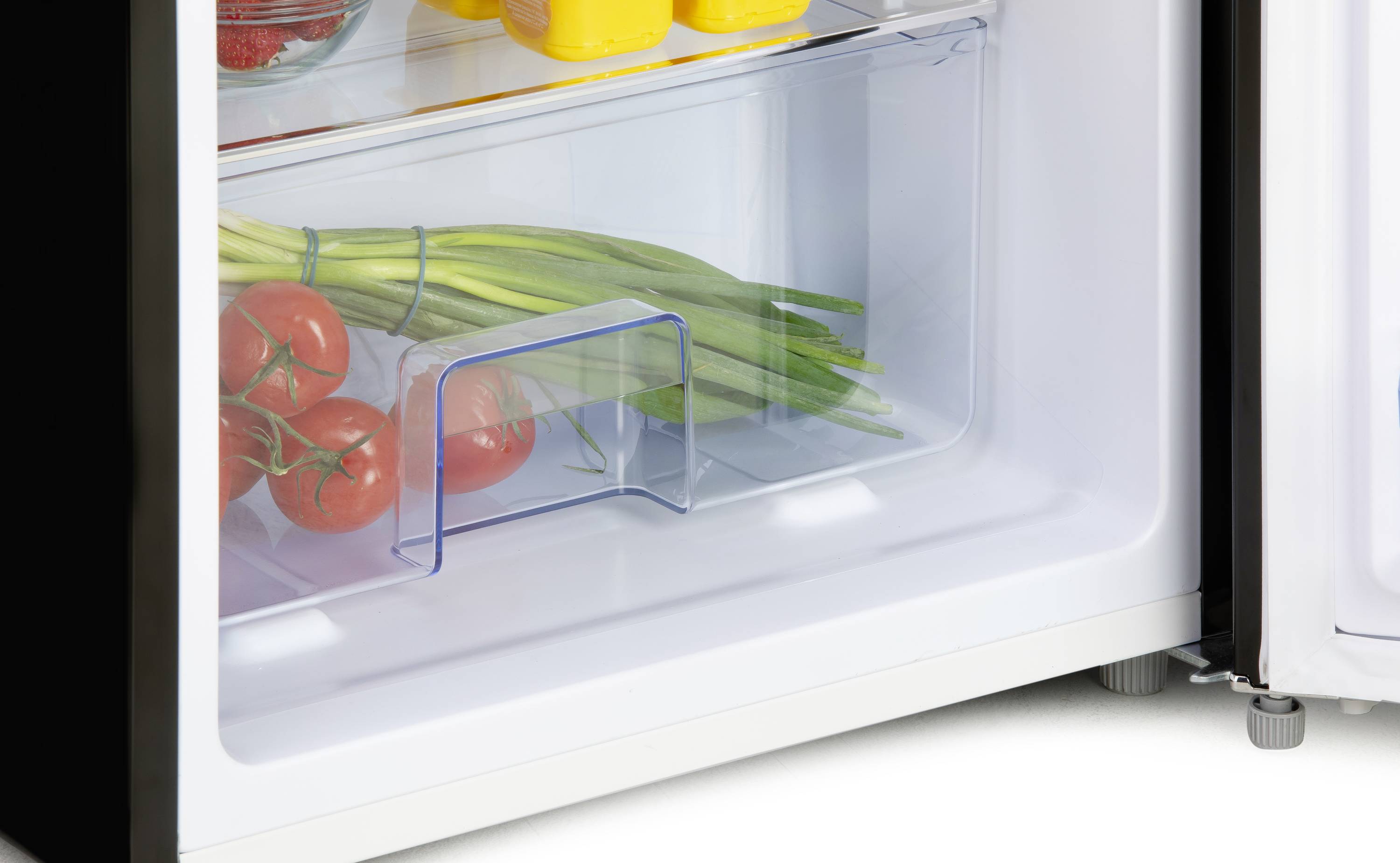 Un réfrigérateur ouvert avec des tomates et du poireau dans le bac inférieur, au-dessus des bouteilles jaunes et un bol de fraises.