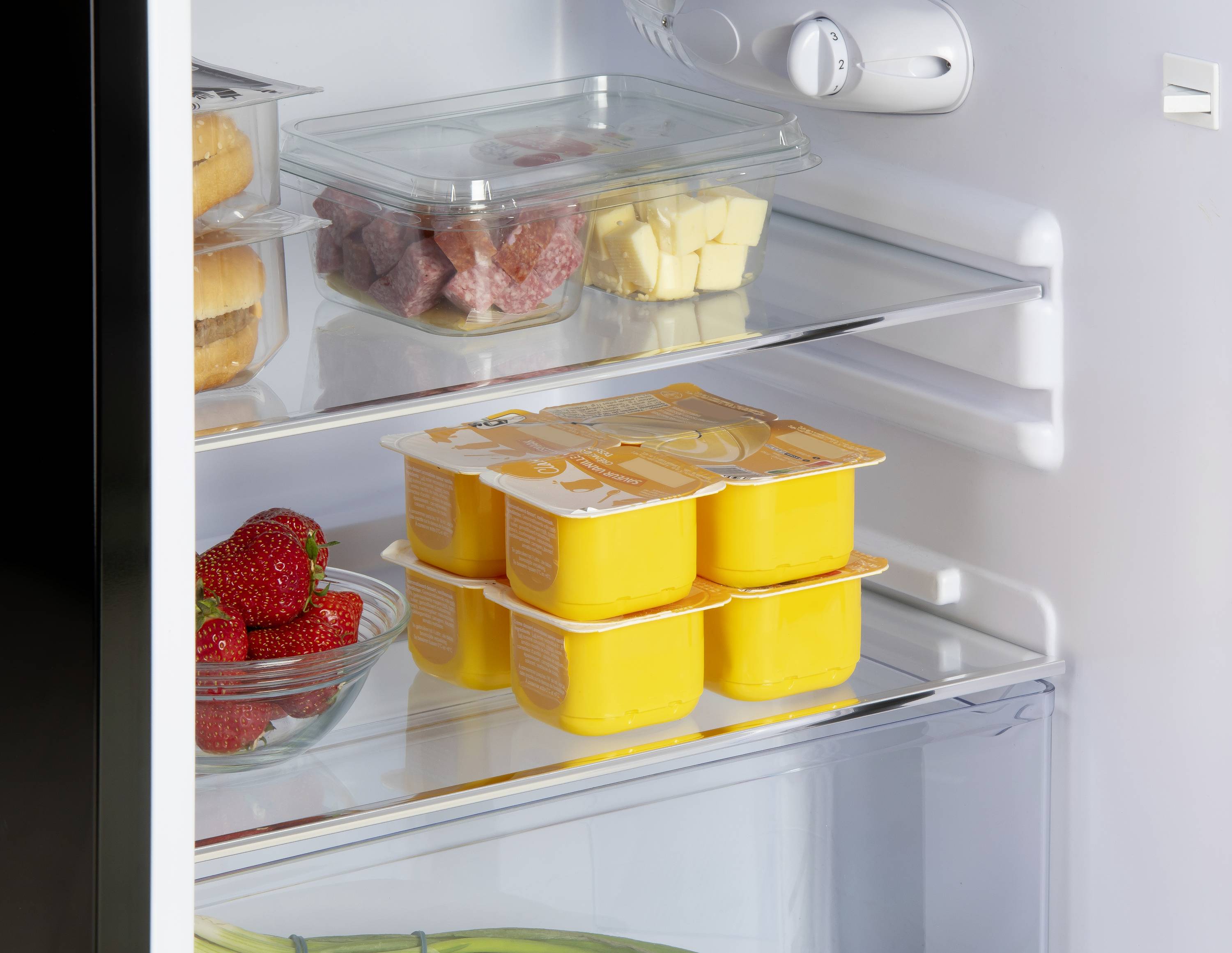 Réfrigérateur avec des étagères en verre ; sur lesquelles sont disposés des saucisses, du fromage, des fraises, des yaourts et des bananes en bas. Les aliments sont soigneusement rangés.