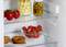 Des tomates, un bol de fraises, des oignons marinés, des olives et deux donuts sont placés sur différentes étagères du réfrigérateur ouvert.