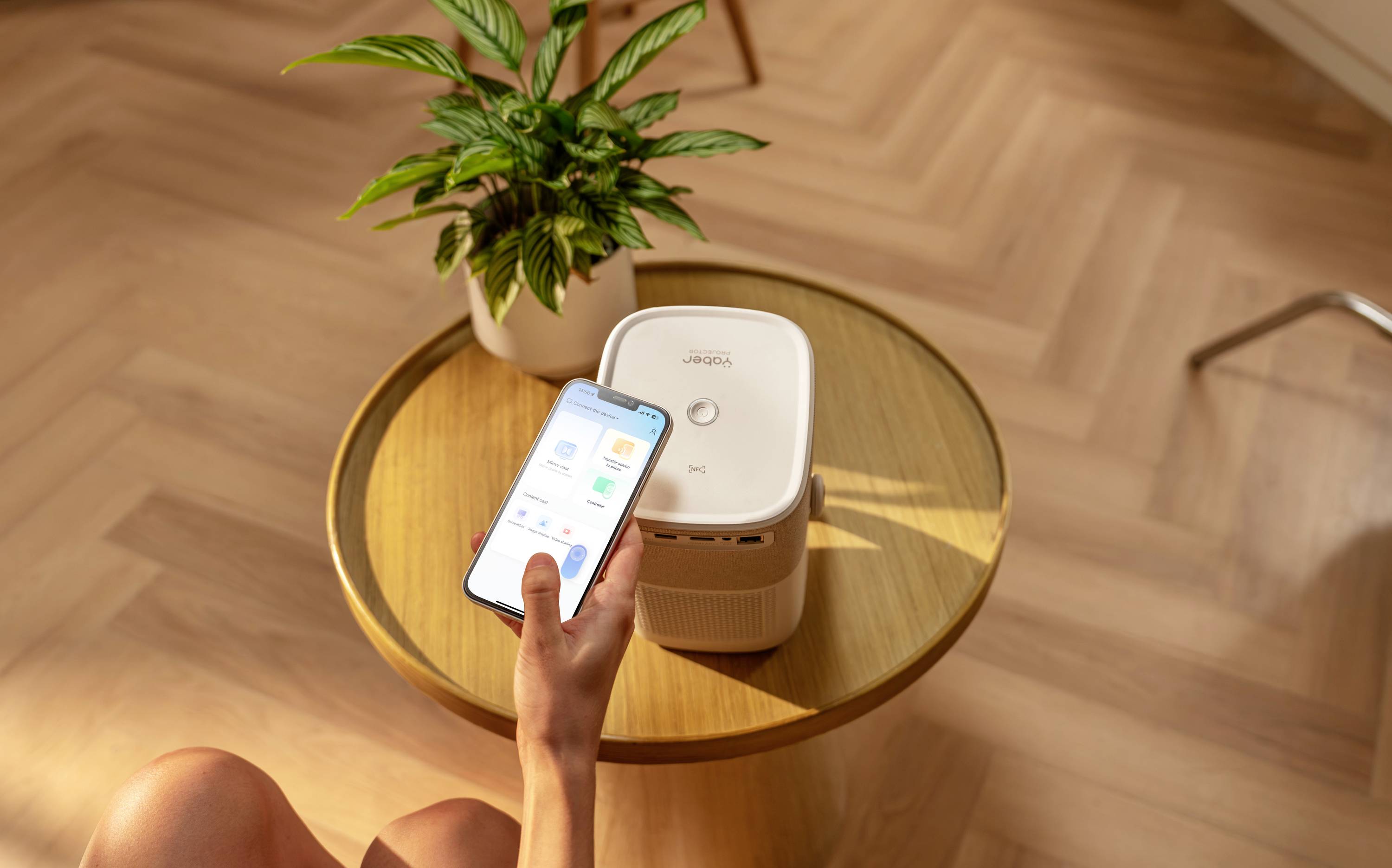 Une personne contrôle un purificateur d'air depuis son smartphone, posé sur une table ronde. À côté de l'appareil se trouve une plante verte.
