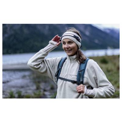 Femme souriante en tenue de randonnée ajuste son bandeau tandis qu'elle fait une randonnée près d'un lac avec des montagnes en arrière-plan.