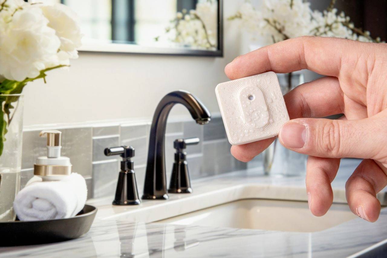 Une main tient un petit capteur carré devant un lavabo de salle de bain moderne, entouré de fleurs et de serviettes.