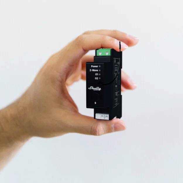 Une main tient un appareil électronique noir et rectangulaire. Sur la face avant de l'appareil sont écrits les mots 'Power' et 'Shelly'.