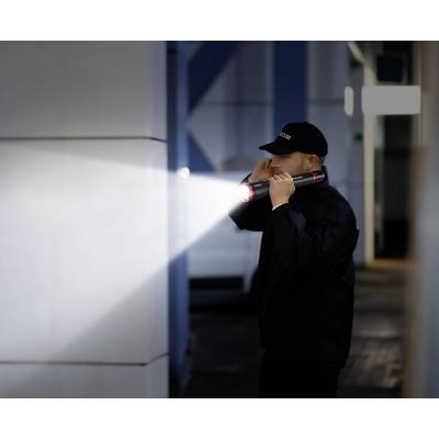 Un agent de sécurité inspecte une zone sombre à l'aide d'une lampe torche. Il porte un uniforme et une casquette, l'environnement ressemble à un site industriel.