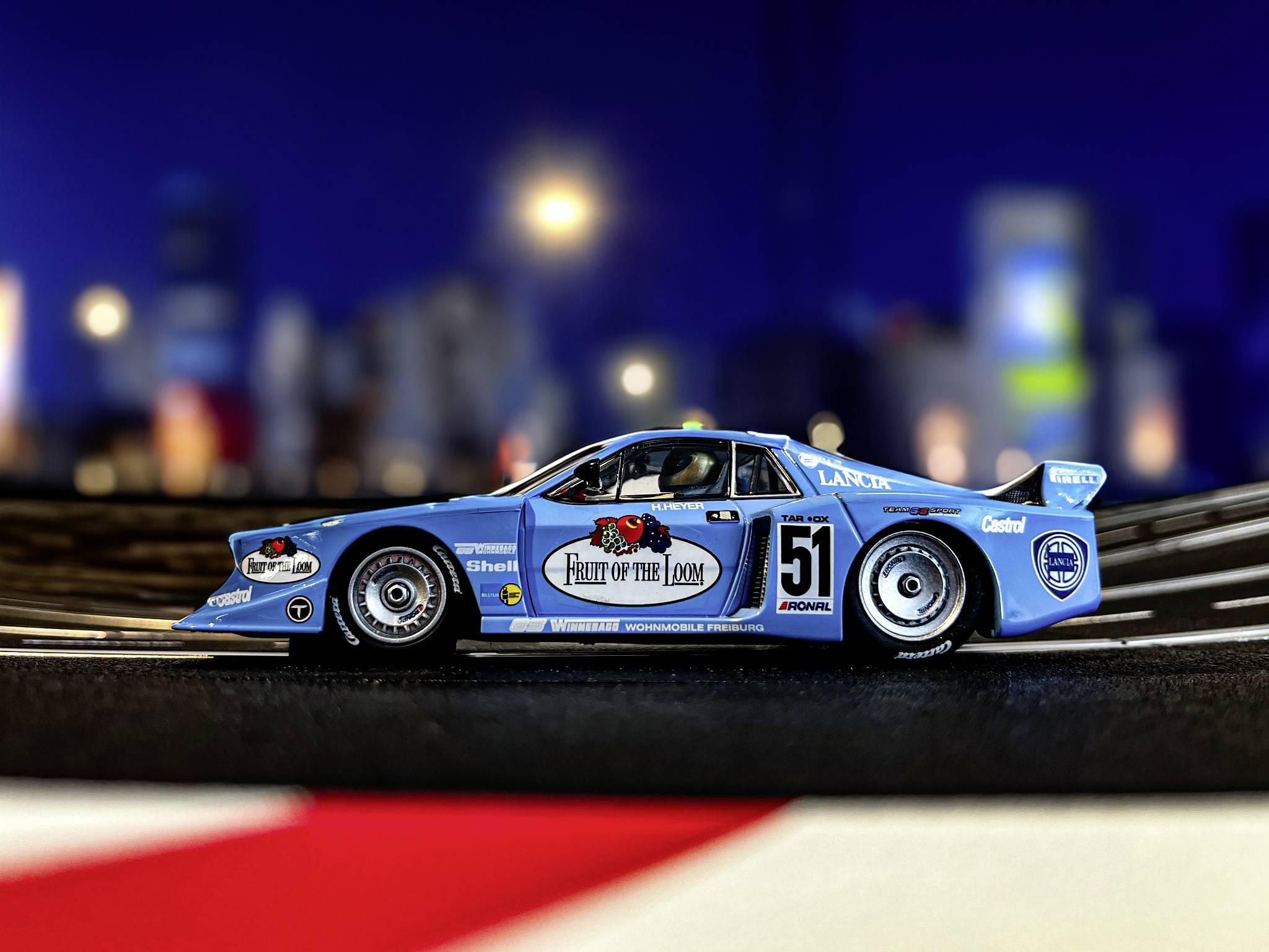 Une voiture de course avec une peinture bleue, portant le numéro 51 et divers autocollants de sponsors, roule sur un circuit de course la nuit.