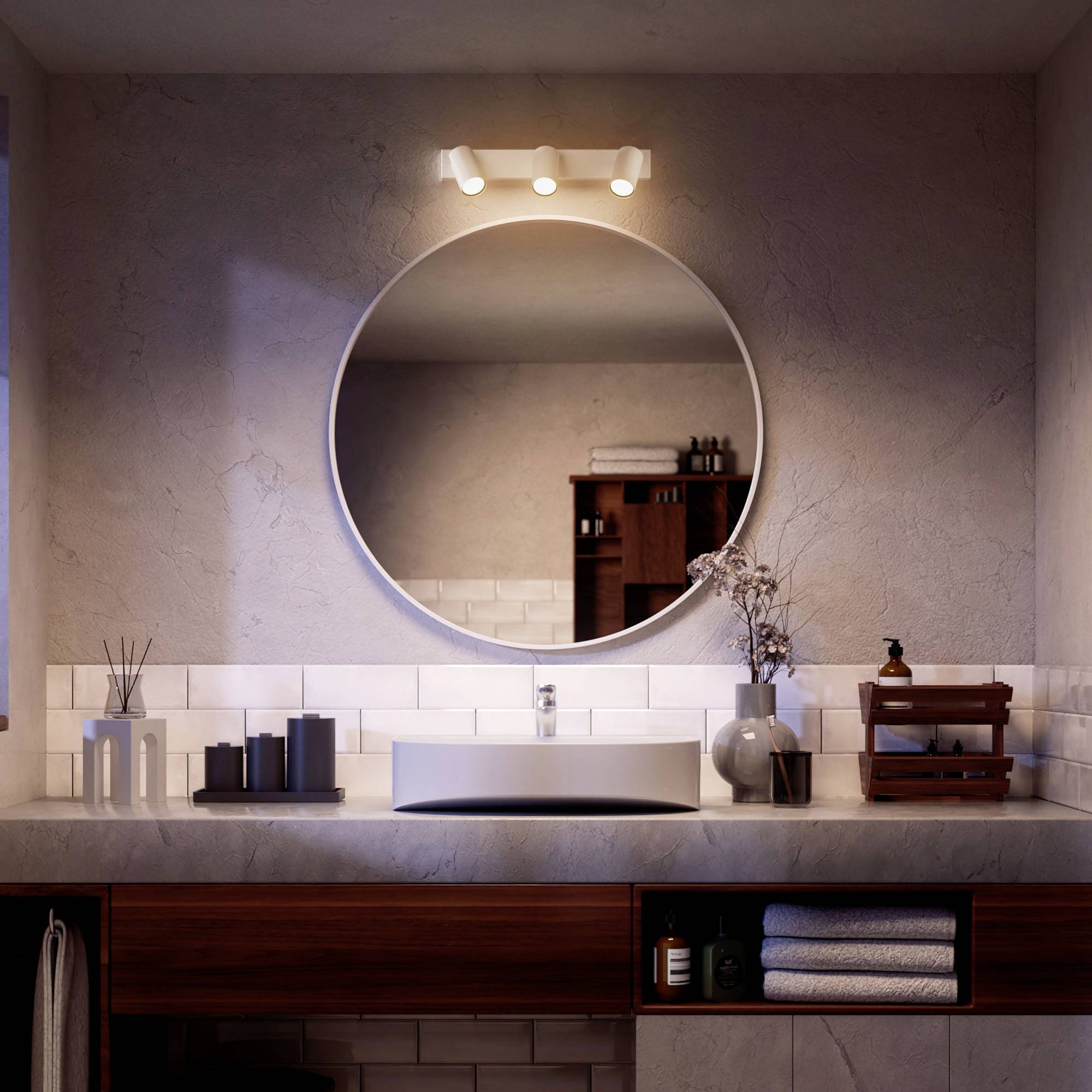 Une salle de bain avec un miroir rond au-dessus d'un lavabo. Un vase de fleurs et une lampe sont posés sur le meuble-lavabo.