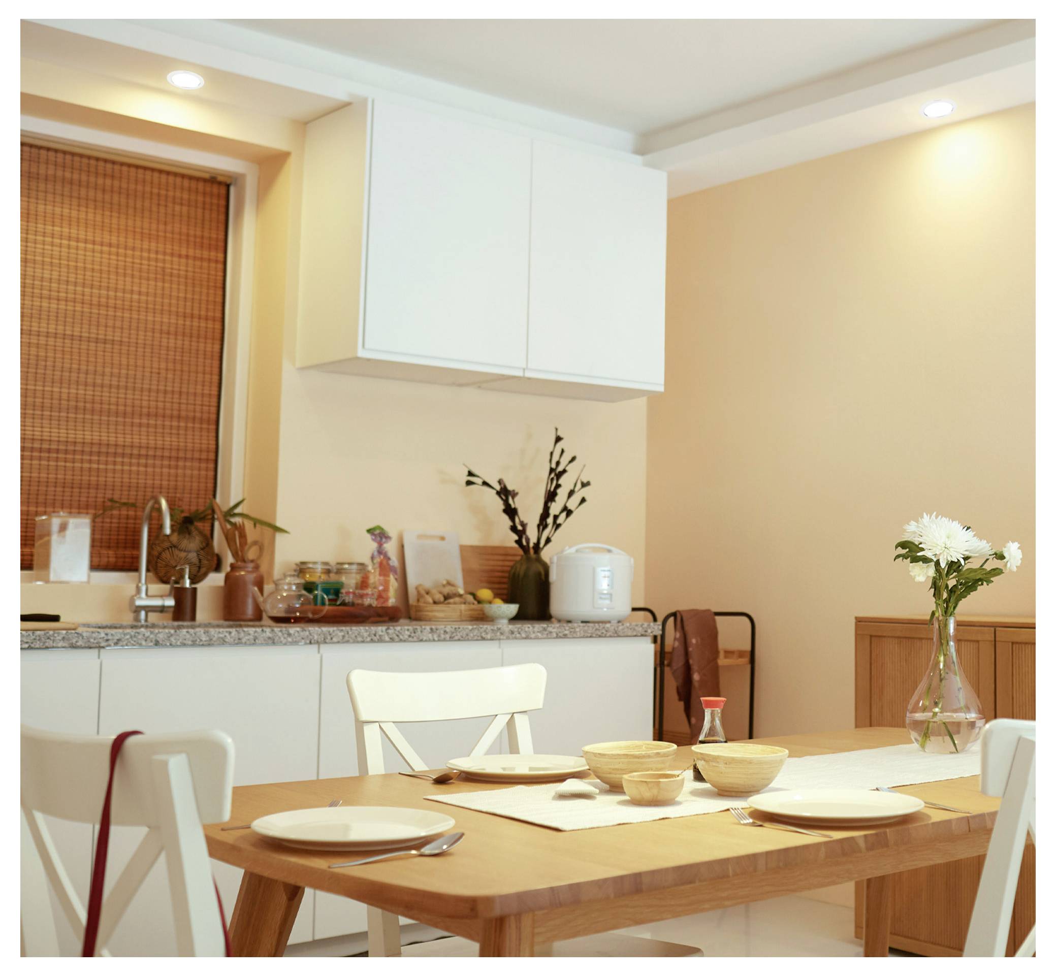 Zone de repas lumineuse avec une table en bois dressée pour un repas, des chaises blanches, un comptoir en granit, des armoires et un vase de fleurs dans le coin.