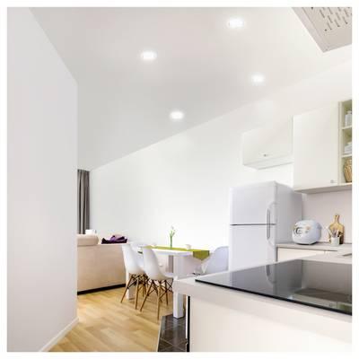 Appartement moderne lumineux en open-plan avec îlot de cuisine, table à manger, chaises blanches, canapé et réfrigérateur sous des luminaires encastrés.