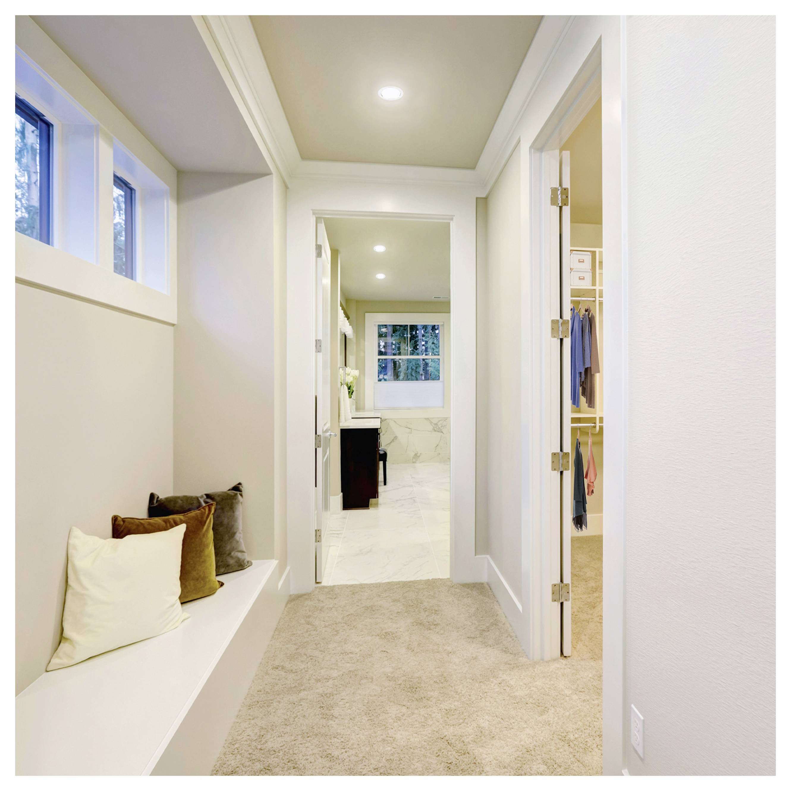Couloir lumineux avec un banc-fenêtre intégré et des coussins, menant à une salle de bain et un dressing, avec des luminaires encastrés au plafond et une moquette beige.