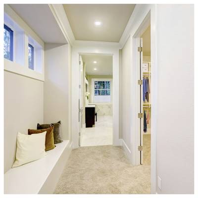 Couloir lumineux avec un banc-fenêtre intégré et des coussins, menant à une salle de bain et un dressing, avec des luminaires encastrés au plafond et une moquette beige.