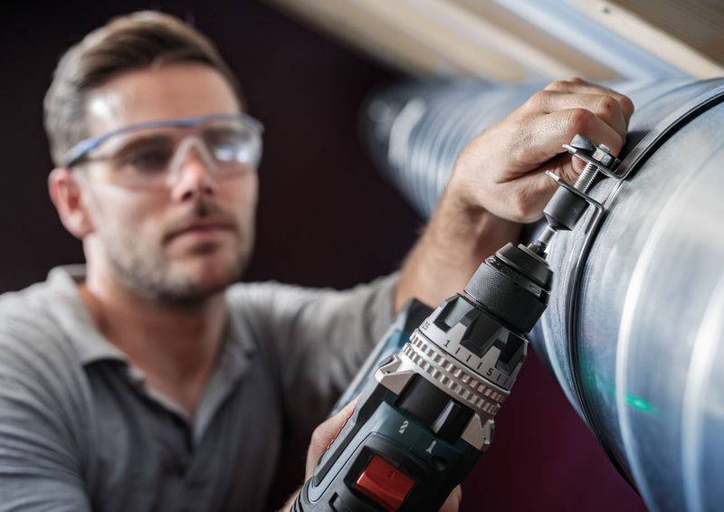 Une personne portant des lunettes de protection installe un grand tube métallique à l'aide d'une visseuse dans un environnement industriel.