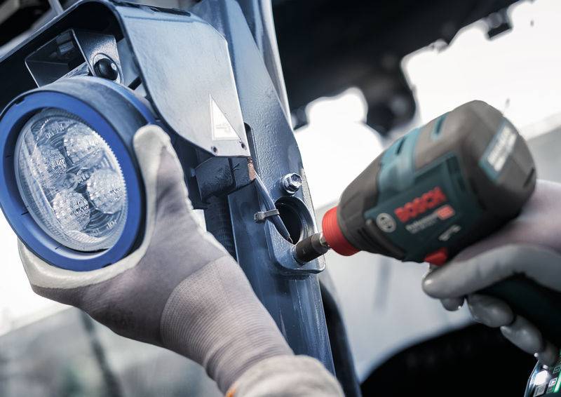 Une personne installe un projecteur LED circulaire sur un support métallique à l'aide d'une visseuse sans fil. Des vêtements de travail et des gants sont visibles.