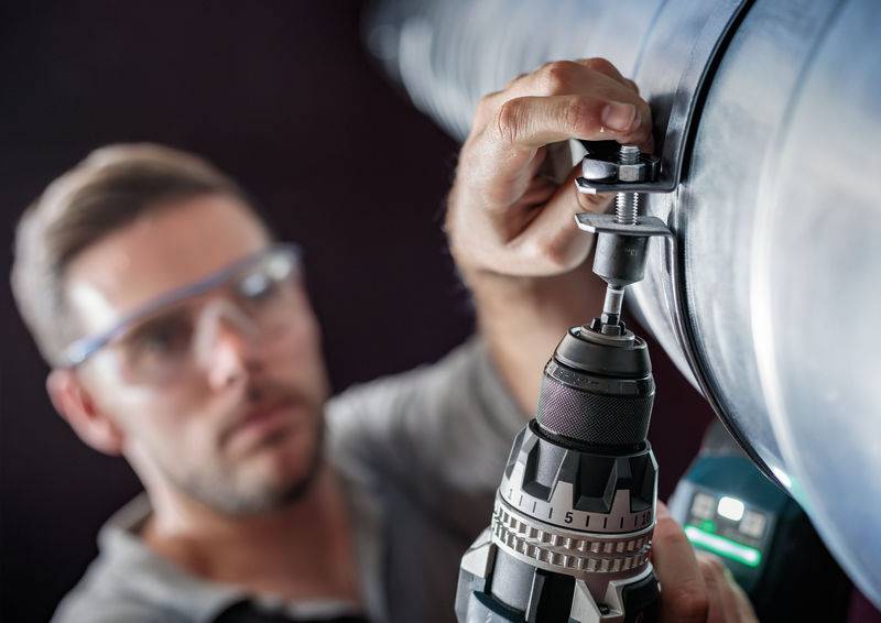 Un homme porte des lunettes de protection et utilise une perceuse pour travailler sur un grand tuyau métallique. Le focus est mis sur l'outil et la main.
