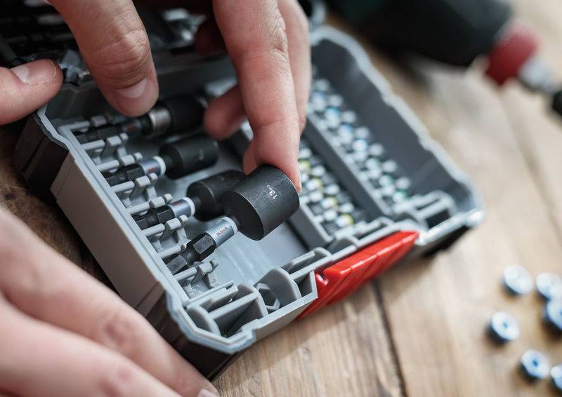 Une personne place une pièce d'outillage dans une boîte comportant plusieurs compartiments, posée sur une table en bois. L'accent est mis sur le rangement et l'organisation des outils.