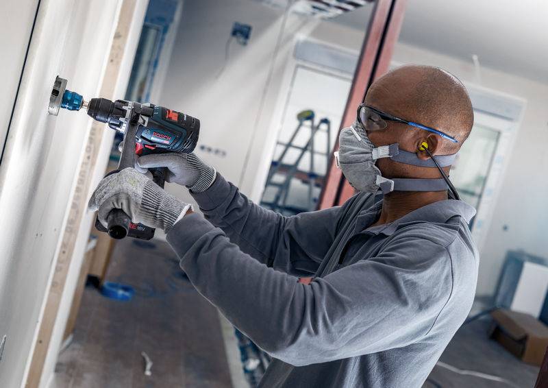Un artisan équipé d'un équipement de sécurité fore un mur dans une pièce en cours de construction. Des échelles sont visibles en arrière-plan.