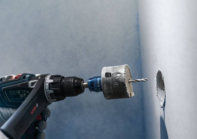 Une perceuse avec un embout est utilisée pour percer un trou rond dans un mur clair. Un trou préexistant est visible.