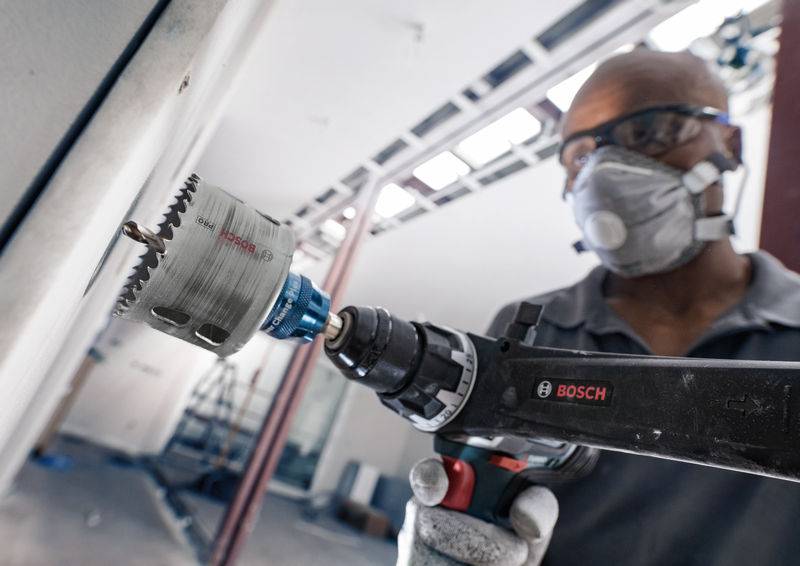 Un ouvrier fore un trou dans un mur à l'aide d'une perceuse Bosch. Il porte des lunettes de protection et un masque. En arrière-plan, un environnement de chantier.