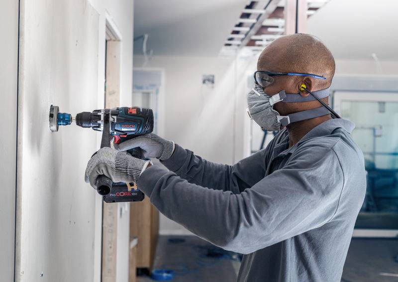 Un ouvrier porte des lunettes de protection et un masque et utilise une perceuse pour faire un trou dans un mur intérieur.