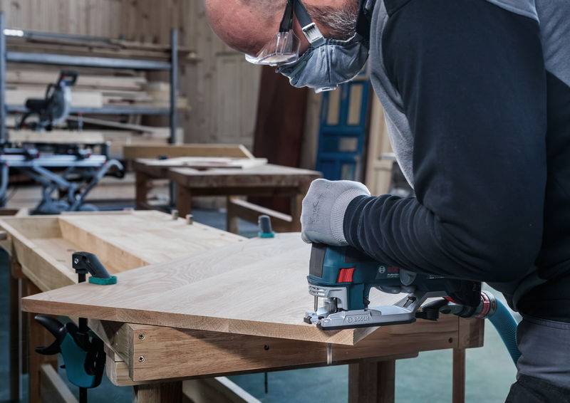 Un homme portant des lunettes de protection et un masque scie du bois avec une scie sauteuse électrique dans un atelier. À l'arrière-plan, on aperçoit d'autres planches de bois.