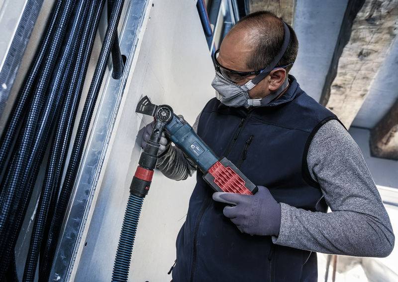 Une personne porte des vêtements de protection et utilise un outil électrique pour travailler sur un mur. Elle porte des lunettes de protection et un masque.