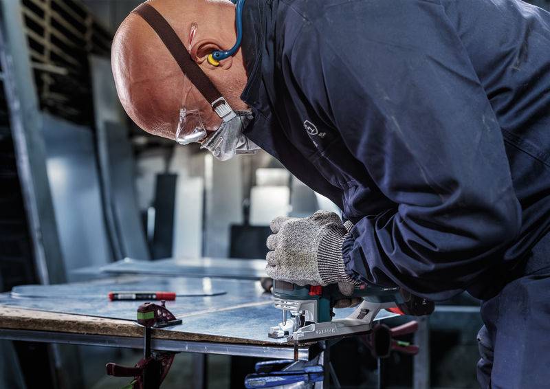 Une personne portant des lunettes de protection et des protections auditives travaille avec une scie électrique portative sur une pièce métallique dans un environnement d'atelier.