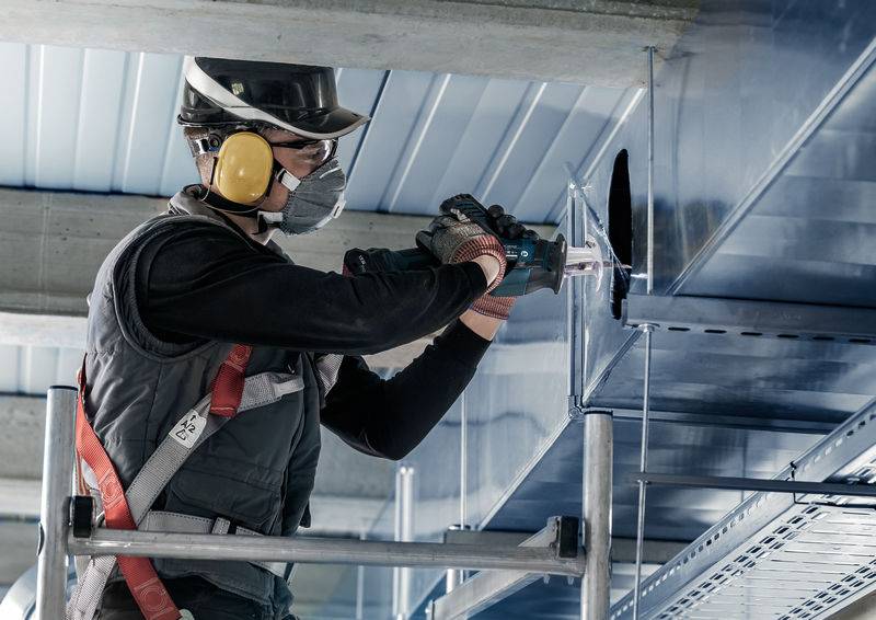 Un ouvrier, portant son équipement de sécurité, fixe un conduit de ventilation à l'aide d'une perceuse dans un bâtiment.