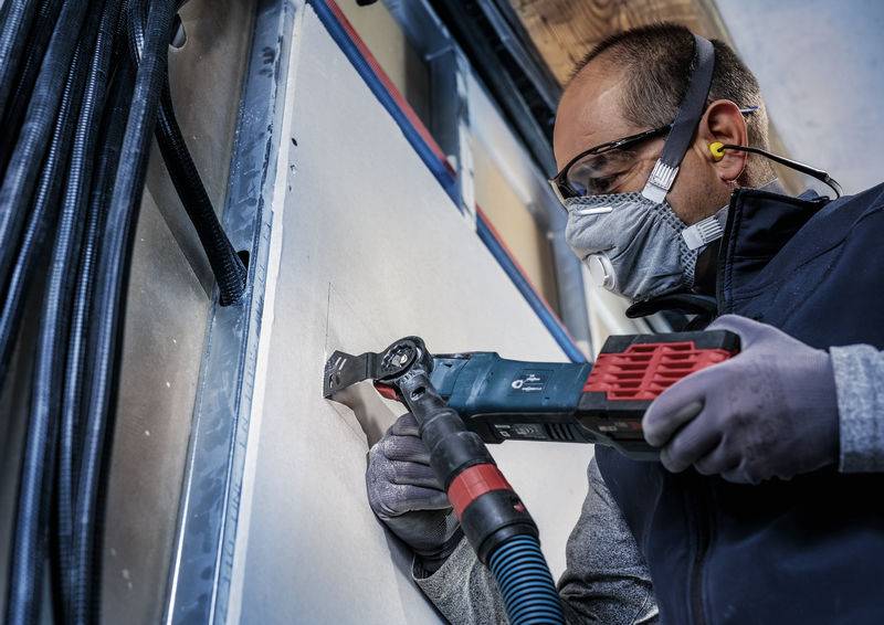 Une personne portant des lunettes de protection et un masque utilise un outil électrique pour travailler une plaque ou un mur.