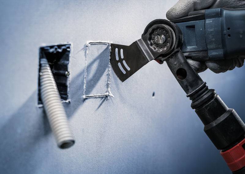 Un artisan utilise un outil multifonction électrique pour découper une ouverture dans un mur, à proximité d'un câble.
