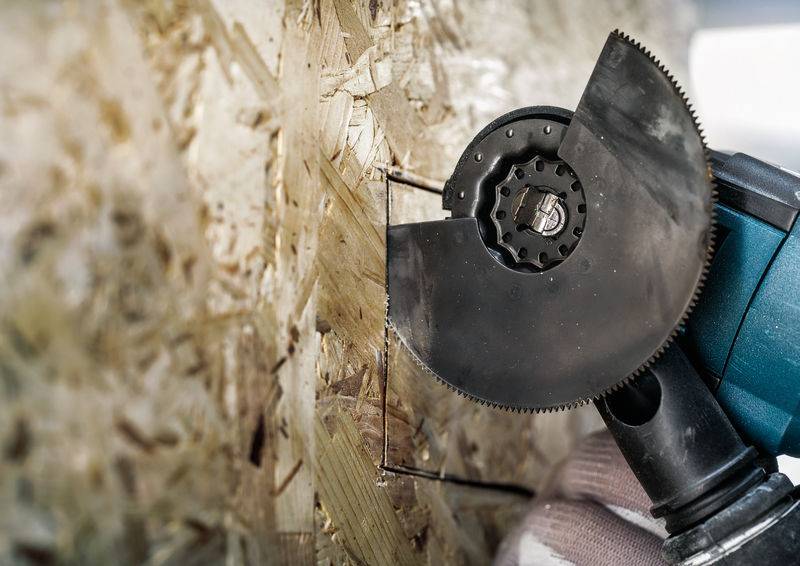 Une personne coupe un matériau dur, probablement du bois ou du panneau de particules, à l'aide d'une meuleuse d'angle. L'outil et la structure sont visibles.