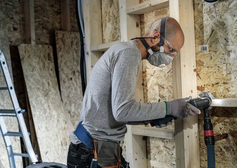 Une personne portant des lunettes de protection et un masque ponçe du bois dans un environnement d'atelier. Des planches de bois et une échelle sont visibles en arrière-plan.