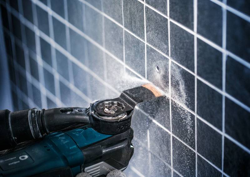 Une personne utilise un outil électrique pour découper un trou carré dans un carrelage mural. De la poussière est visible.