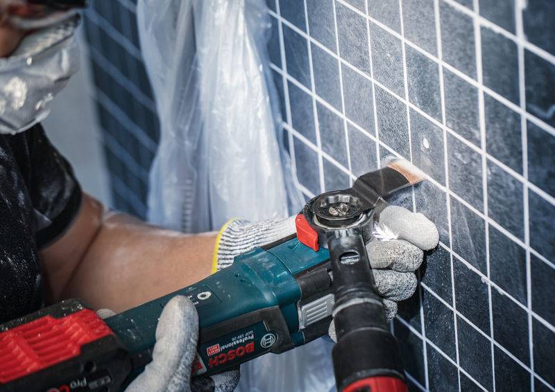 Une personne utilise un outil électrique pour travailler des carreaux sur un mur. Des lunettes de protection et des gants sont portés.